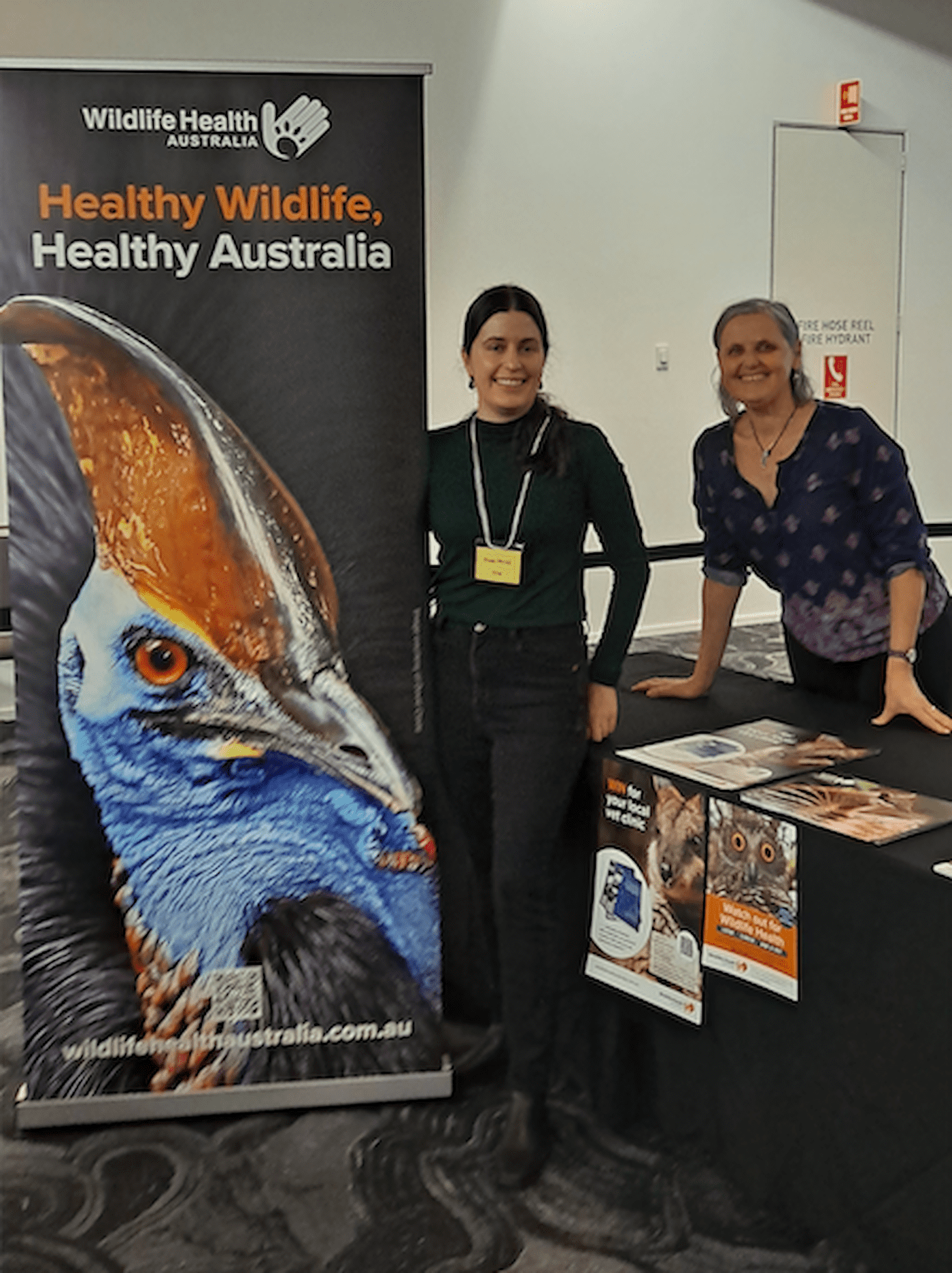 Simone Vitali and Shana Ahmed at a conference, standing in front of a exhibition table and next to a pull up banner with an image of a cassowary. 