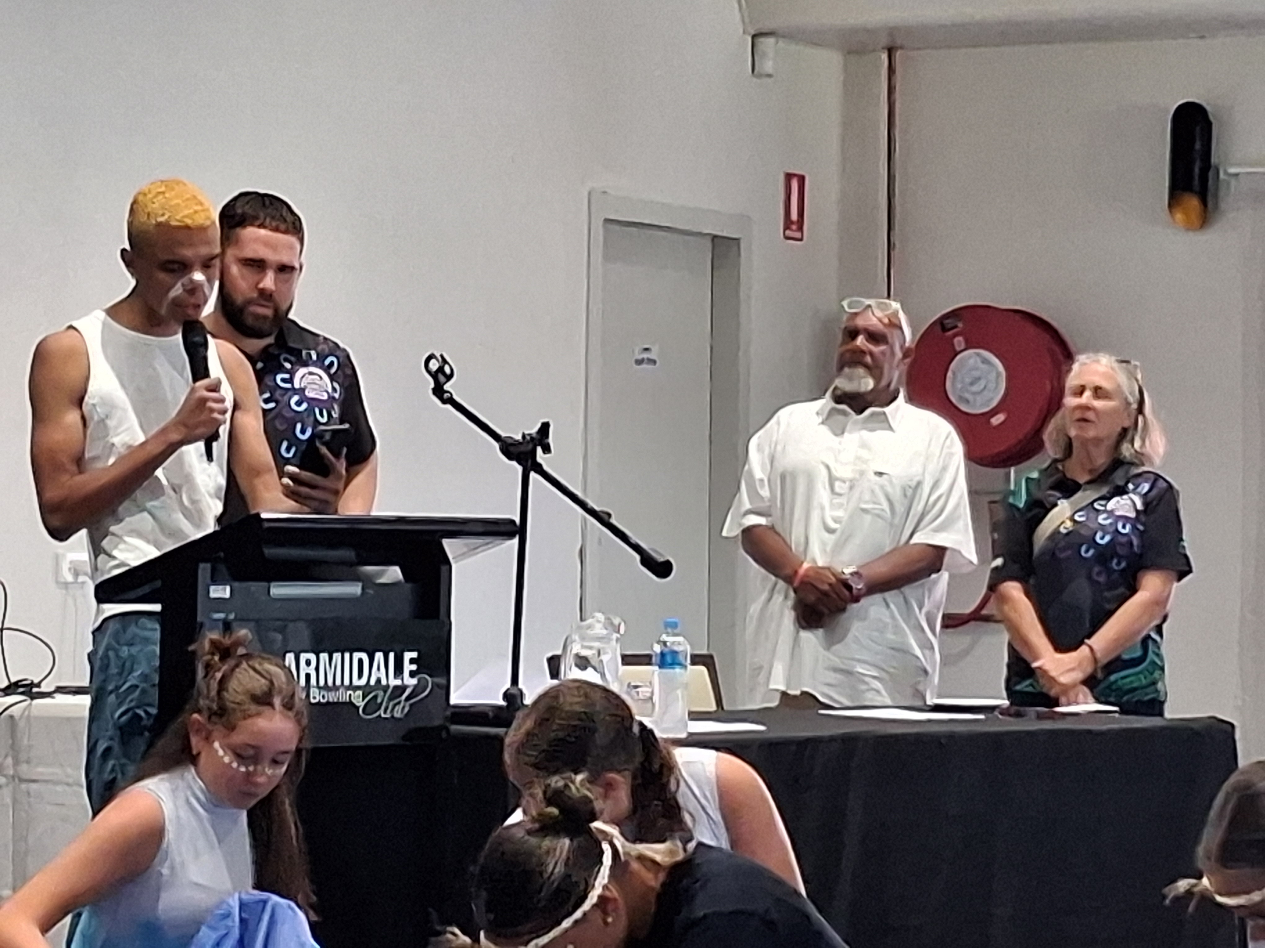 First Nations speaker with a microphone at a podium, with 2 men and 1 women watching on. 